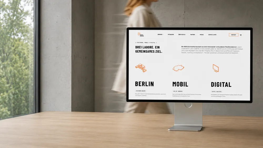 A computer monitor on a desk displaying a website with the words 'DREI LABORE. EIN GEMEINSAMES ZIEL.'. A person is blurred in the background walking past a window. The screen shows sections titled 'BERLIN,' 'MOBIL,' and 'DIGITAL.'