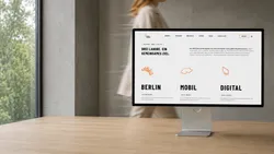 A computer monitor on a desk displaying a website with the words 'DREI LABORE. EIN GEMEINSAMES ZIEL.'. A person is blurred in the background walking past a window. The screen shows sections titled 'BERLIN,' 'MOBIL,' and 'DIGITAL.'