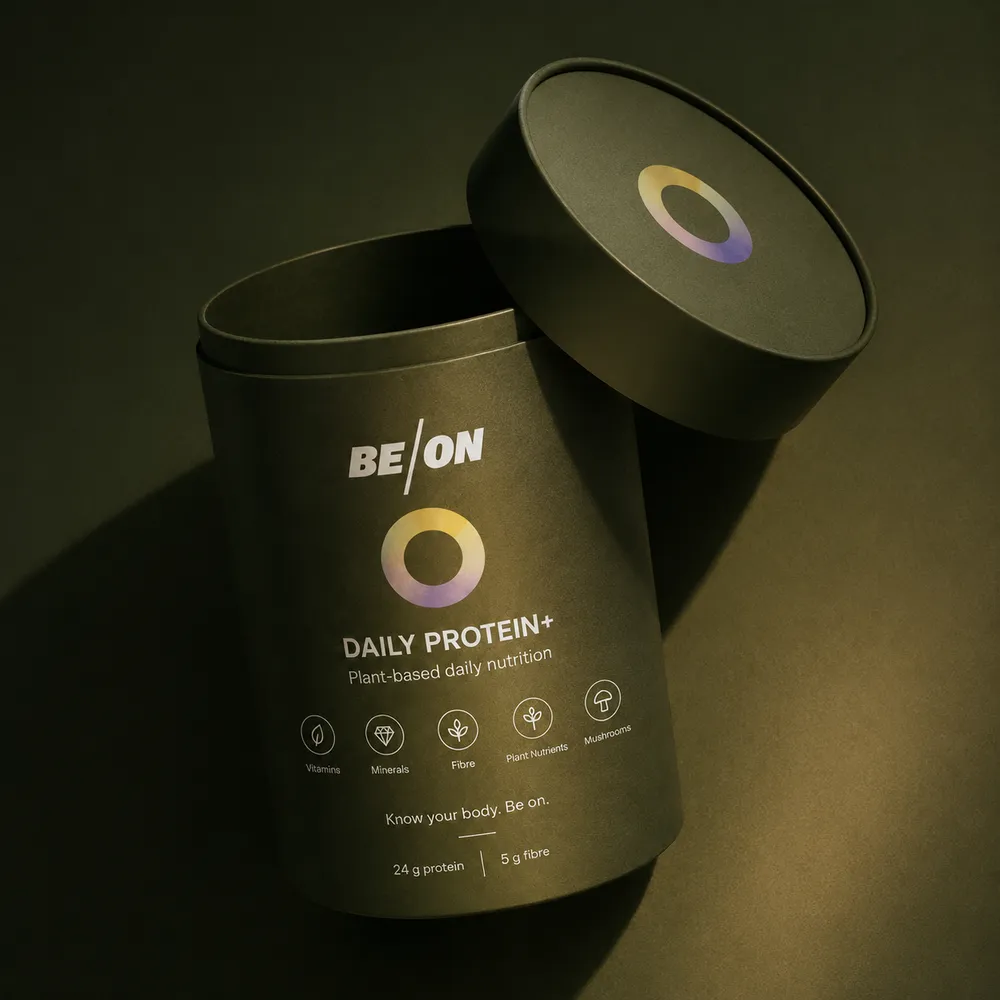 A cylindrical black package of BE/ON Daily Protein+ with a partially open lid. The label indicates it is plant-based nutrition, featuring an abstract circular logo and icons for vitamins, minerals, fiber, plant nutrients, and mushrooms. It states '24 g protein' and '5 g fibre.'