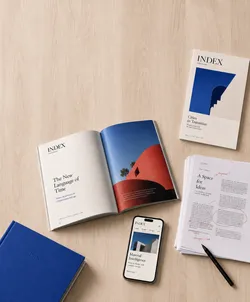 A flat lay of design materials and a smartphone on a wooden surface. The items include an open magazine displaying an article titled 'The New Language of Time,' with a large photo of a red wall against a blue sky. An adjacent pile of marked-up papers, a black pen, a blue book, and a smartphone showing an article titled 'Material Intelligence' are also visible.
