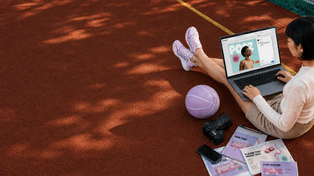 A person sitting on a red sports court with a laptop showing a colorful design, surrounded by a lavender basketball, camera, smartphone, and design pamphlets.