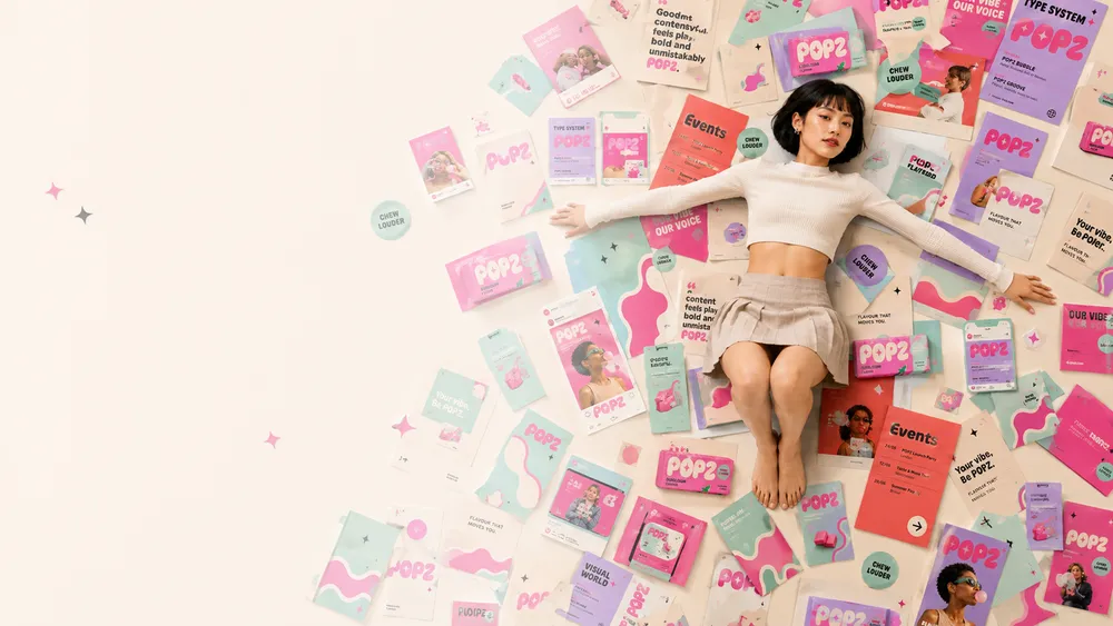 A woman with short dark hair lies on a floor covered with colorful posters and packages labeled 'POP2'. She wears a cropped white sweater and a beige skirt, with her arms and legs extended. The materials around her are predominantly pink, purple, and teal, featuring various texts and graphics.