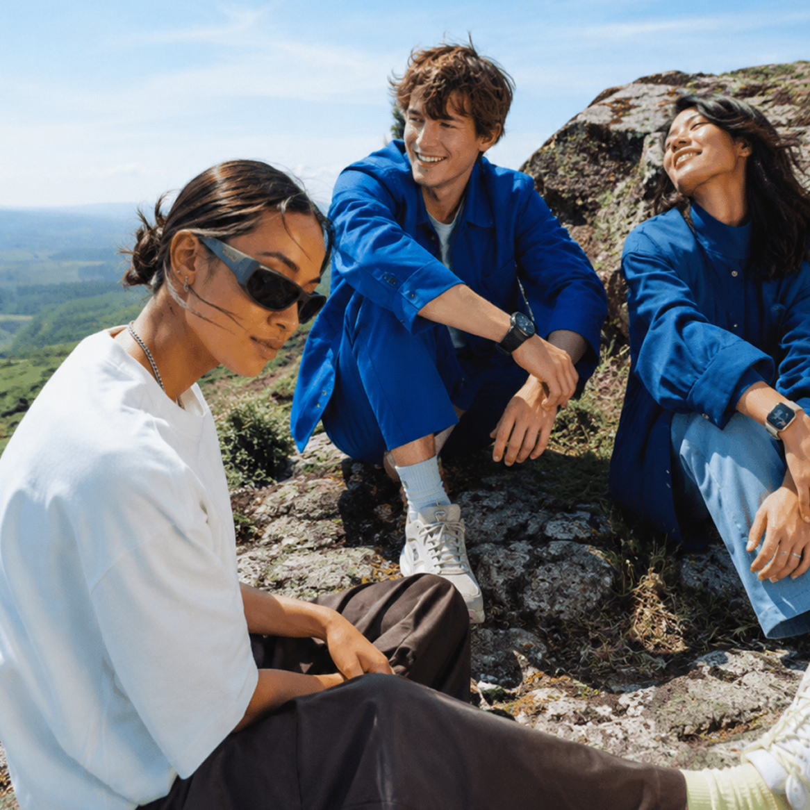 Drei Personen sitzen lächelnd auf einem Felsen in freier Natur. Eine Person trägt eine weiße Bluse und Sonnenbrille, die anderen eine blaue Jacke. Es ist ein sonniger Tag mit blauem Himmel und grünem Tal im Hintergrund.