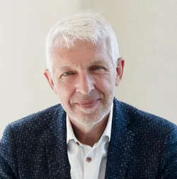 Ein lächelnder Mann mit grauem Haar in einem dunklen, gemusterten Anzug und weißem Hemd, sitzt vor einem neutralen Hintergrund.