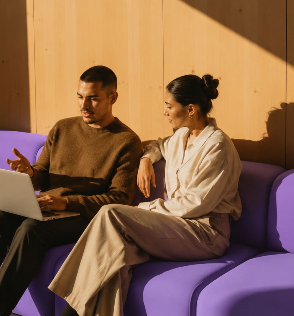 Ein Mann und eine Frau sitzen auf einem lila Sofa und betrachten einen Laptop. Der Mann trägt einen braunen Pullover, während die Frau ein helles Hemd und weite Hosen trägt. Sie sitzen vor einer Holzvertäfelung und sind im Sonnenlicht.