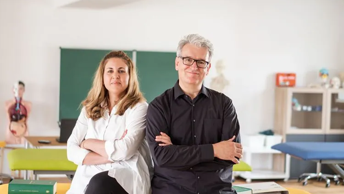 Ein Mann und eine Frau sitzen nebeneinander auf einem Tisch in einem Klassenzimmer. Beide tragen lässige Kleidung, die Frau hat dunkles langes Haar, der Mann hat graues Haar und trägt eine Brille. Im Hintergrund befinden sich ein anatomisches Modell, ein Schwarzes Brett und Regale.