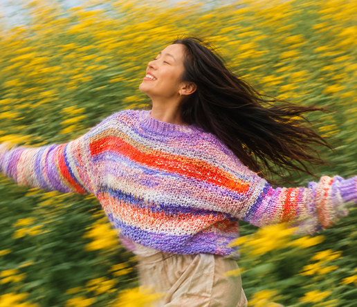 Eine Frau mit einem bunten gestrickten Pullover genießt die Freiheit in einem Feld voller gelber Blumen. Sie lächelt und breitet die Arme aus, während ihr Haar im Wind weht.