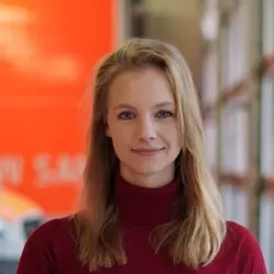 Eine Frau mit langen blonden Haaren trägt einen roten Rollkragenpullover und steht vor einem verschwommenen Hintergrund mit orangefarbenen und grauen Elementen.