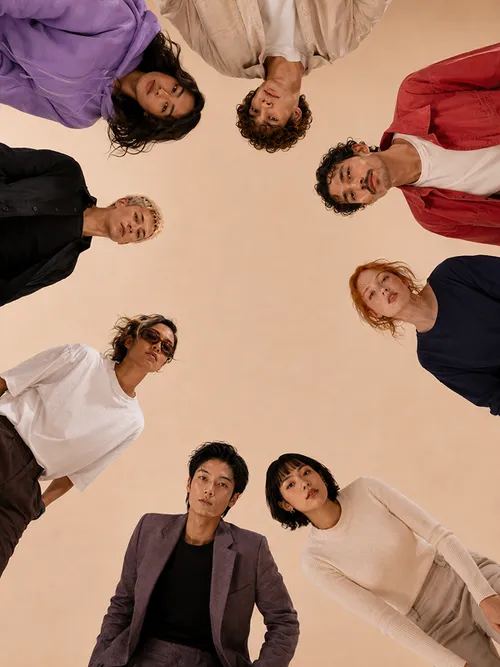 A group of eight people stands in a circle and looks down into the camera while wearing various casual outfits. The background is beige and the perspective is from below, positioning them all around the edge of the image.
