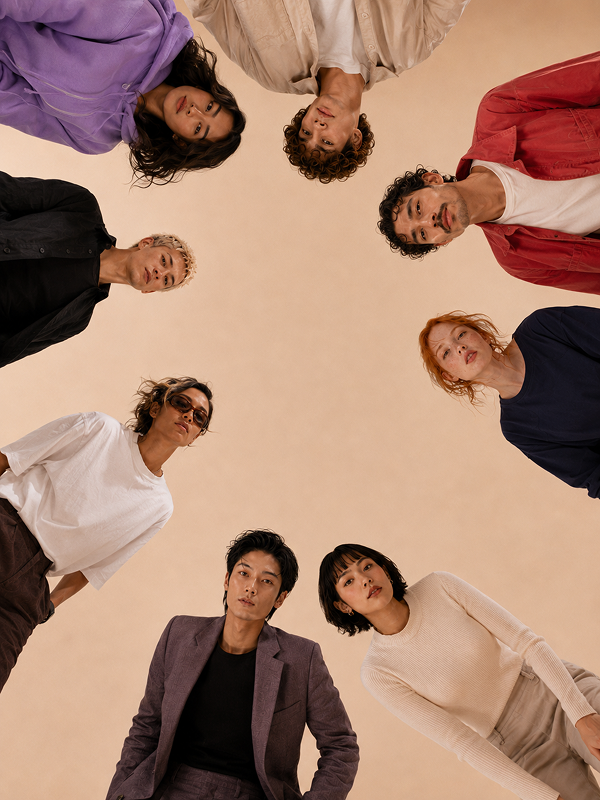 Eine Gruppe von acht Personen steht im Kreis und blickt nach unten in die Kamera, während sie verschiedene lässige Outfits tragen. Der Hintergrund ist beige und die Perspektive ist von unten, wodurch sie alle um den Bildrand herum positioniert sind.