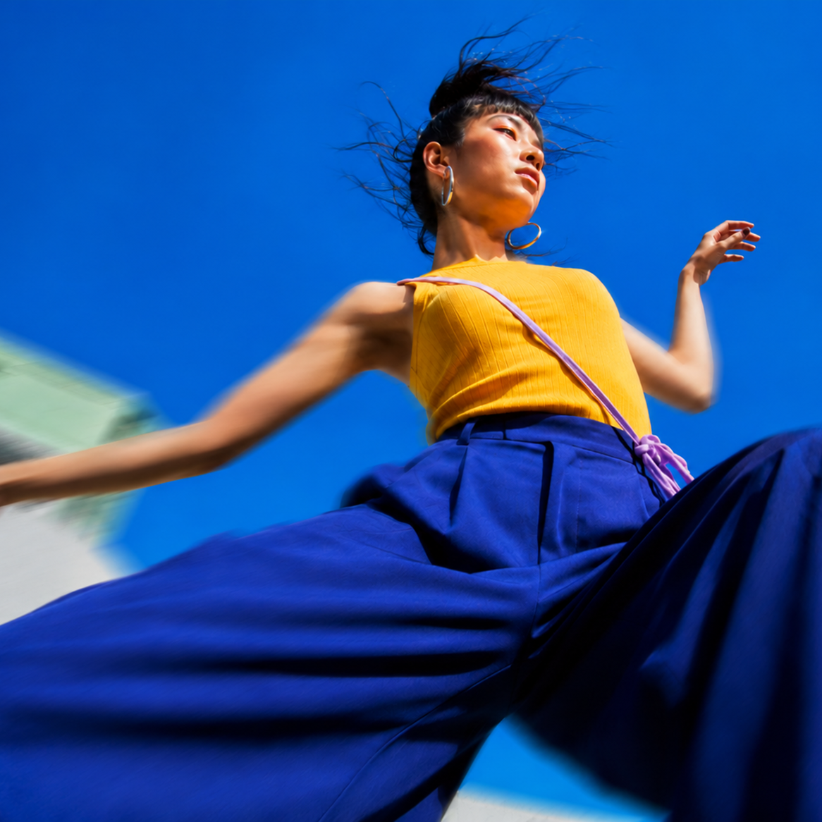 Frau in gelbem Oberteil und blauer Hose vor blauem Himmel, in bewegter Pose.