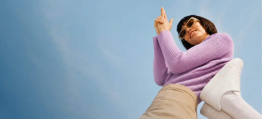 Woman in purple sweater and sunglasses, photographed from below. She is holding her hands together with fingers pointing upwards while smiling, wearing bright pants and white sneakers.