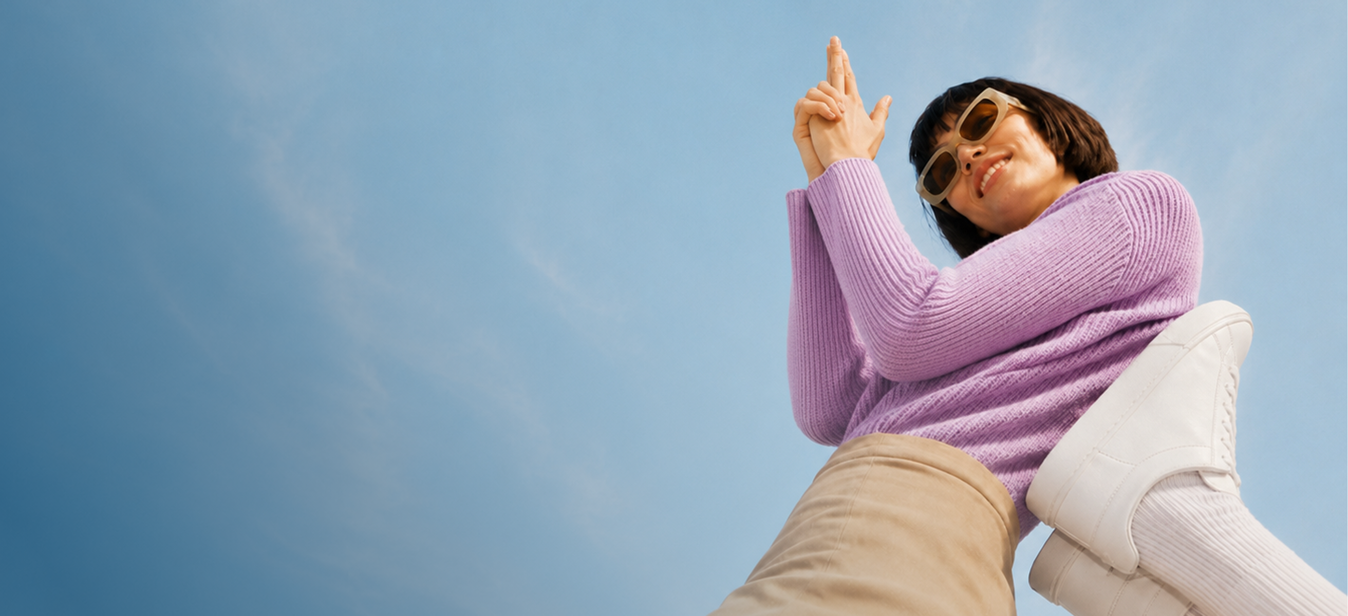 Frau in lila Pullover und Sonnenbrille, von unten fotografiert. Sie hält ihre Hände zusammen, die Finger zeigen nach oben, während sie lächelt und eine helle Hose und weiße Sneaker trägt.