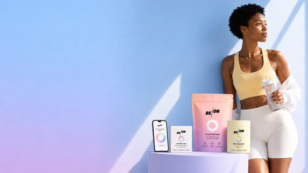 Woman in sportswear holding a water bottle stands next to a table with BE/ON brand products and a smartphone displaying a BE/ON app. The background is a gradient of light blue and pink.