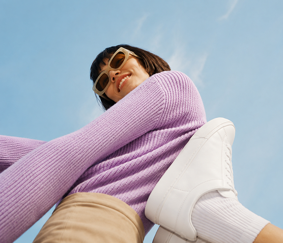 Frau mit Sonnenbrille in lila Pullover und weißem Sneaker von unten fotografiert vor blauem Himmel