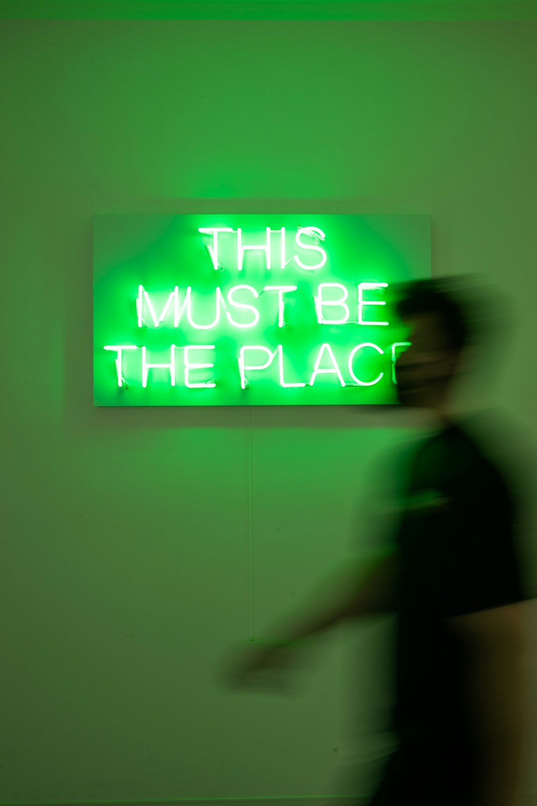 Green neon sign reading 'THIS MUST BE THE PLACE' with a blurred person walking by.