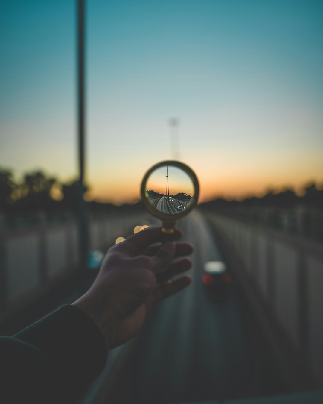 Hand holding a magnifying glass focusing on a road at sunset.