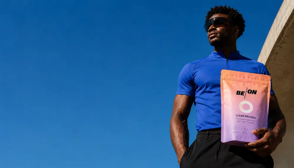 Man in blue shirt and sunglasses holding a bag of BE ON clear protein against a blue sky.