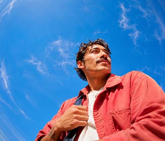 Mann in roter Jacke, der in den Himmel schaut, umgeben von blauen Himmel mit wenigen Wolken. Er hält einen Riemen seiner Tasche. Fotografie aus Untersicht.