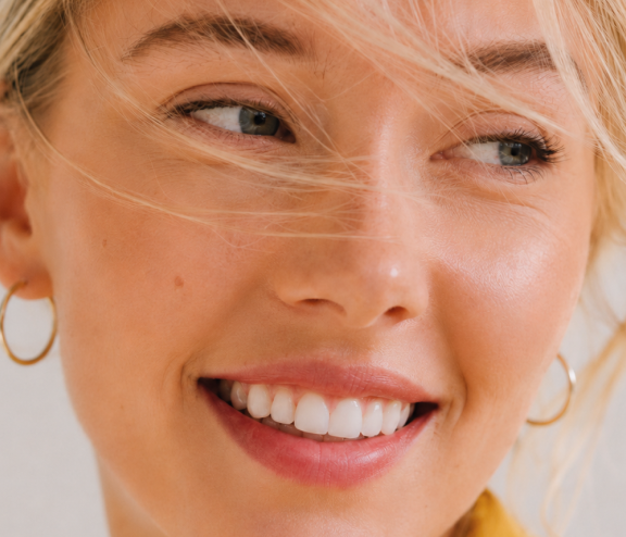 Nahaufnahme einer lächelnden Frau mit blonden Haaren und goldenen Ohrringen. Sie hat helle Haut und ihre Haare wehen leicht im Gesicht.