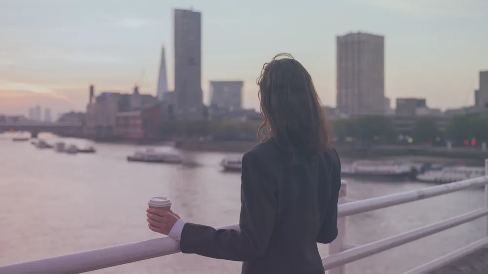 Person holding a coffee cup overlooking a river and city skyline at sunset.