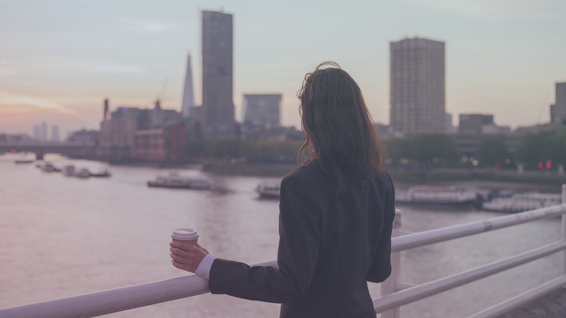 Person holding a coffee cup overlooking a river and city skyline at sunset.