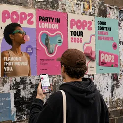 Person holding a phone, looking at a wall with colorful posters advertising 'POPZ' events and products, including a launch party in London and a 2026 tour.