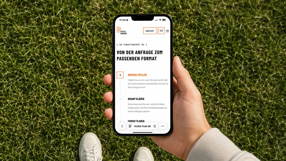 Person holding a smartphone displaying a website in German with the title 'Von der Anfrage zum passenden Format' while standing on grass.