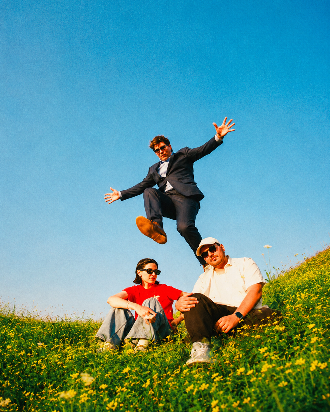 Person in a suit jumps energetically over two seated individuals on a grassy hill with a clear blue sky background. The seated individuals are wearing casual clothing, sunglasses, and seated amidst yellow flowers.