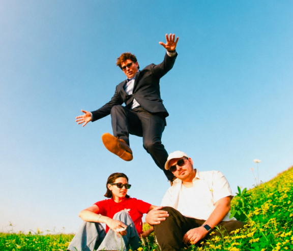 Person in a suit jumps over two seated individuals on a grassy hill with a blue sky background.