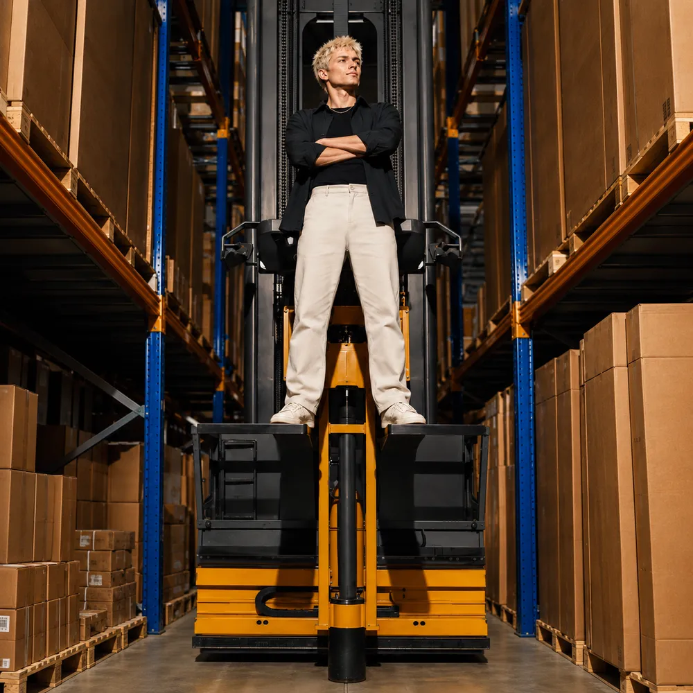 Person standing confidently on a forklift in a warehouse with arms crossed, surrounded by shelves of boxes.