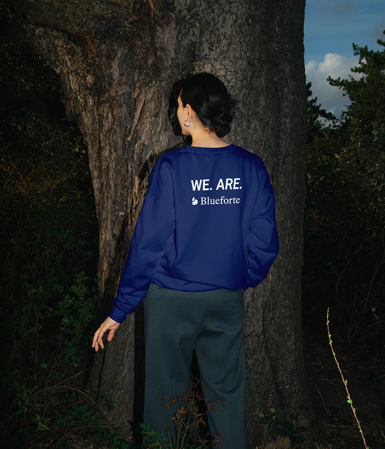 Person standing with back to camera wearing a blue jacket with 'WE. ARE. Blueforte' text, in a wooded area.