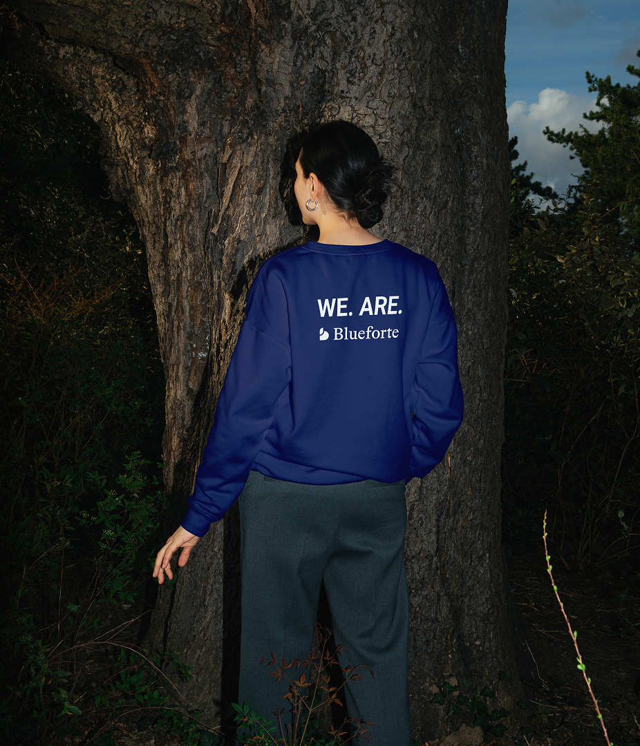 Person standing with back to camera wearing a blue jacket with 'WE. ARE. Blueforte' text, in a wooded area.