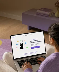 Person using a laptop displaying a webpage for a learning app with a black cat illustration and the text 'Learn smarter. Pass with confidence.' The room has purple decor including a table and books.