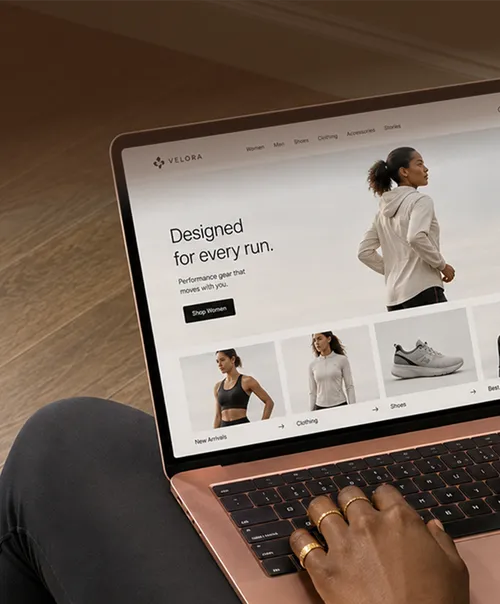 ノートパソコンでスポーツウェアのオンラインストアを閲覧している人です。画面には、'Designed for every run'という文字とスポーツウェアやシューズの画像が表示されています。ノートパソコンはローズゴールドで、この人は指に指輪を着けています。