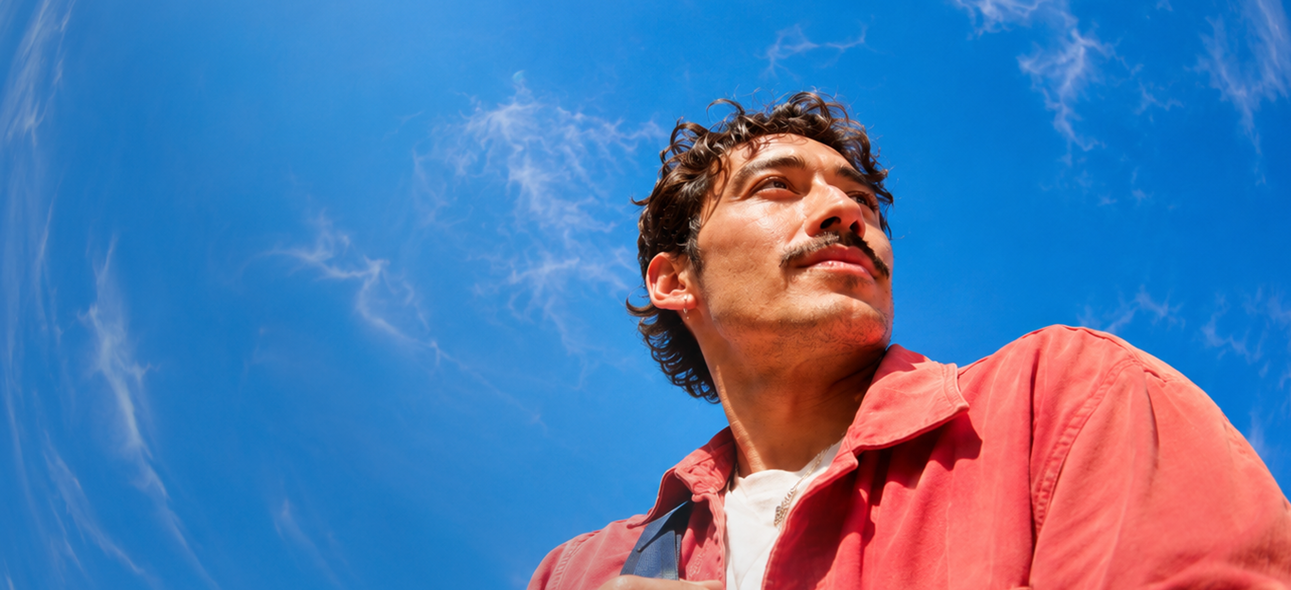 Person wearing a coral jacket looking to the side against a blue sky with wispy clouds.