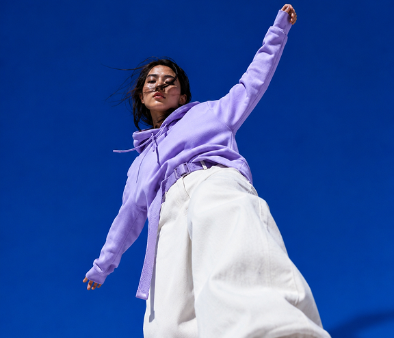 Person wearing a lavender hoodie and white wide-leg pants against a clear blue sky. The perspective is from below, creating a dynamic and dramatic composition.