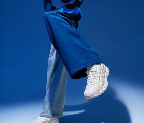 Person wearing white sneakers and blue flared jeans with one leg lifted against a blue background.