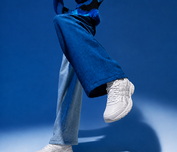 Person wearing white sneakers and blue flared jeans with one leg lifted against a blue background.