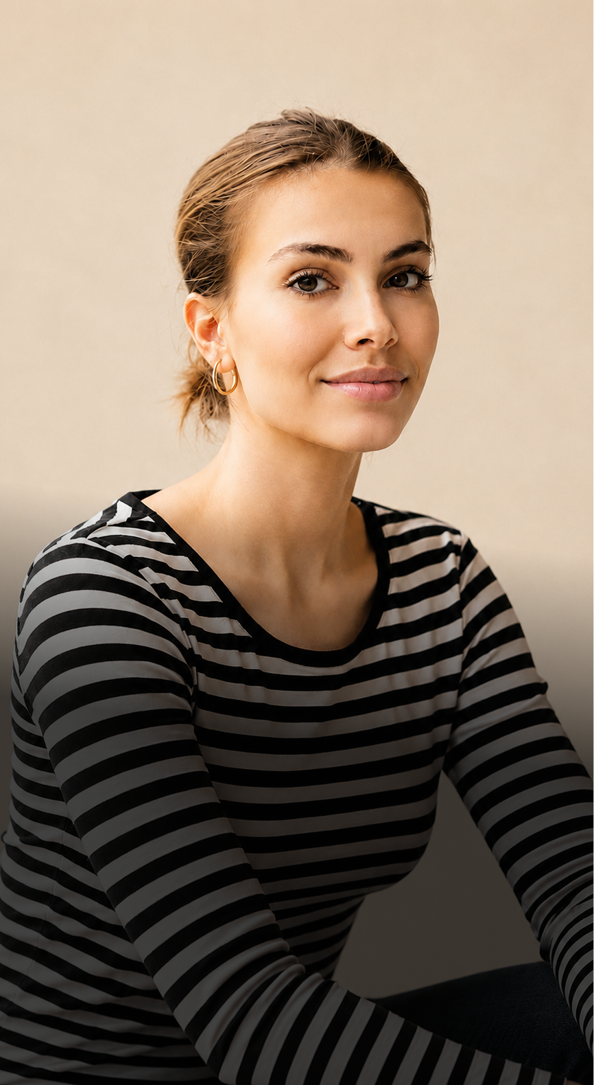 Person with light brown hair tied back, wearing a black and white striped shirt and gold hoop earrings, poses against a beige background.