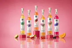 Six bottles of GLOU Vinegary vinegar varieties on a pink background. Each bottle is on a pedestal and has different colors and names: Kräuter Smash, Hamburger Sauer, Rauch Pepper, Streuobstwiese, Hamburger Sauer, and Beere Beere. Pieces of fruit, lime, apple, and orange are lying around them.