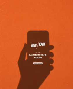 Shadow of a hand holding a smartphone against an orange wall with text promoting a launch for BE/ON, stating 'we're launching soon', and 'stay tuned'.