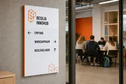 Sign on a wall at Resilia InnoHub displaying directional arrows to Empfang, Workshopraum, and Resilienzlabor. In the background, a group of people is seated at a table in a modern room with orange accents.