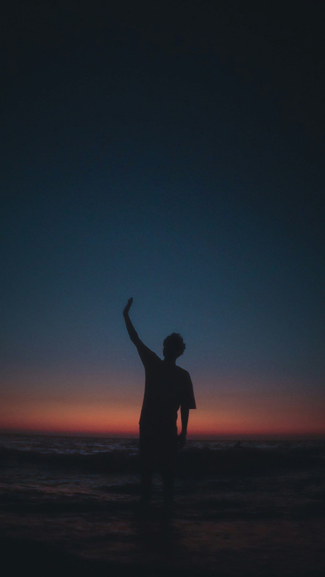 Silhouette of a person raising a hand against a sunset over the ocean.