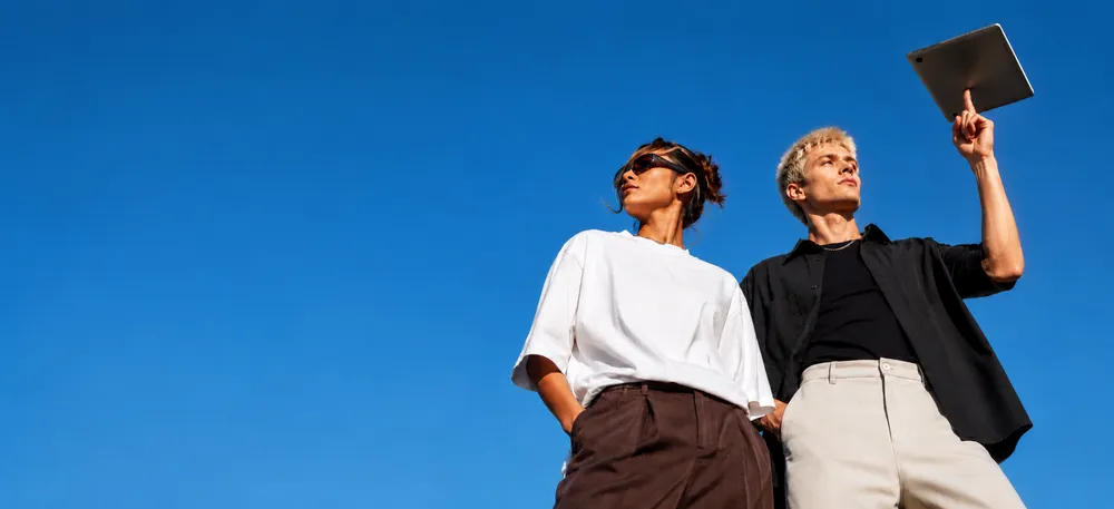 Two individuals standing against a clear blue sky. One person holds a tablet upward, wearing a black shirt and light pants. The other wears a white shirt and dark pants.