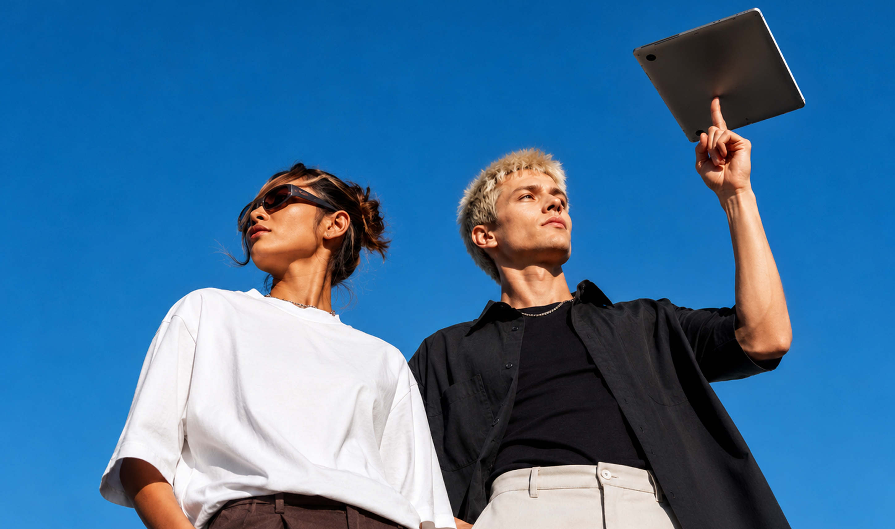 Zwei Personen stehen unter blauem Himmel. Eine Person trägt eine Sonnenbrille und ein weißes T-Shirt, die andere hebt ein Tablet in die Luft und trägt ein schwarzes Hemd über einem dunklen T-Shirt.
