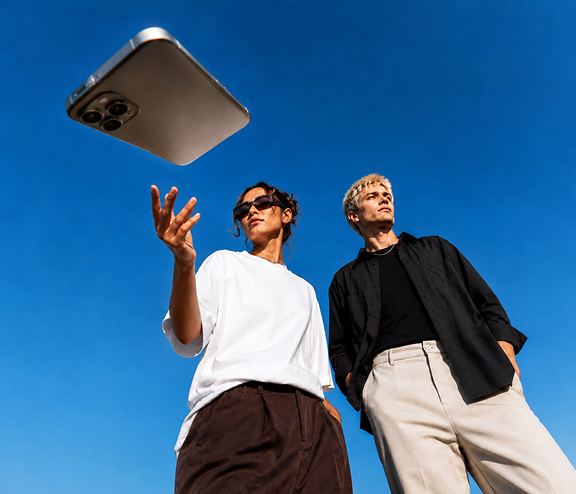 Zwei Personen stehen unter einem klaren blauen Himmel. Eine Person wirft ein Smartphone in die Luft, während die andere daneben steht. Beide tragen lässige Kleidung, eine mit Sonnenbrille. Der Schwerpunkt liegt auf dem in der Luft befindlichen Smartphone.